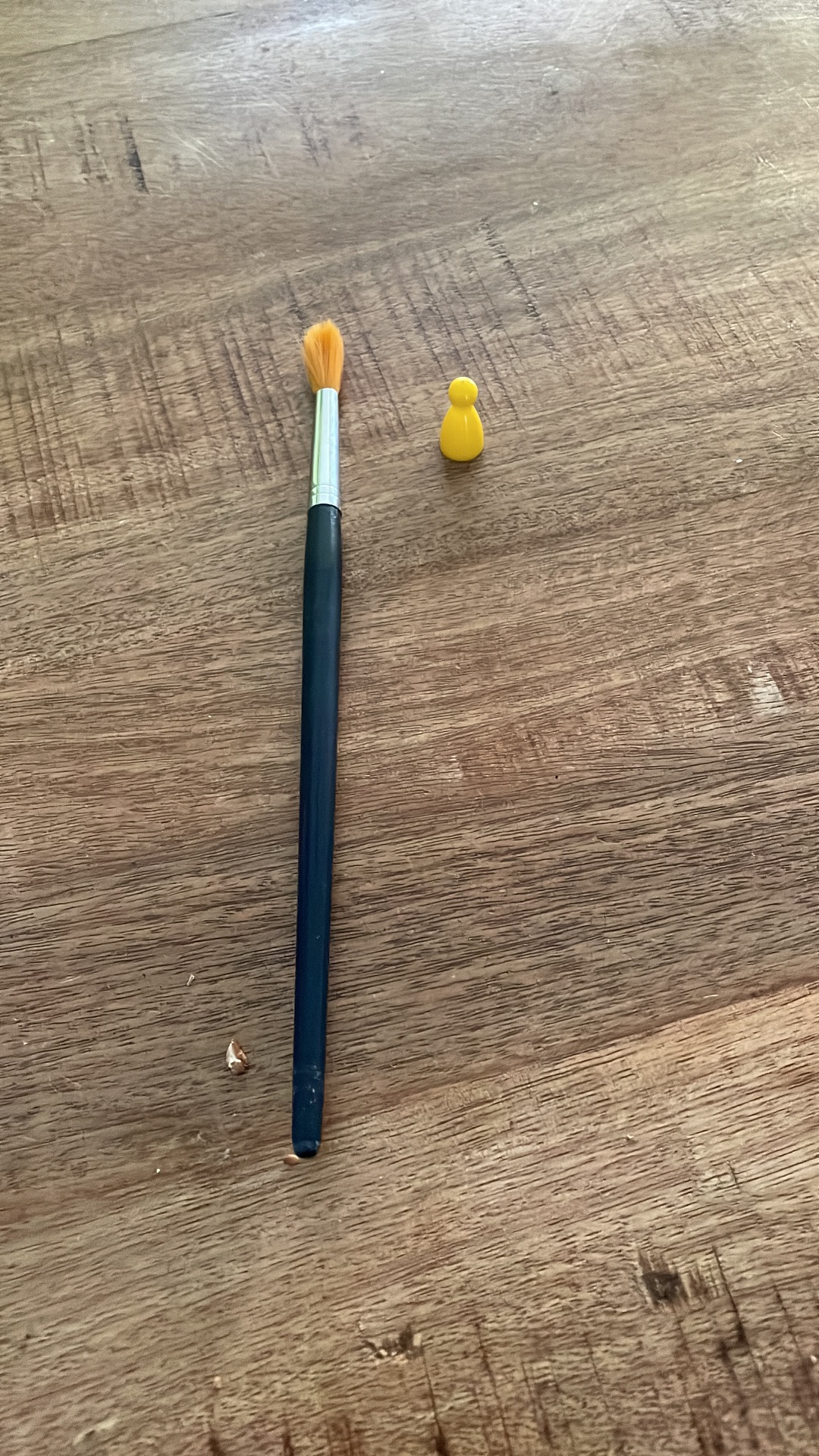 Blauwe verfkwast met gele haren en een klein geel poppetje op houten plank.