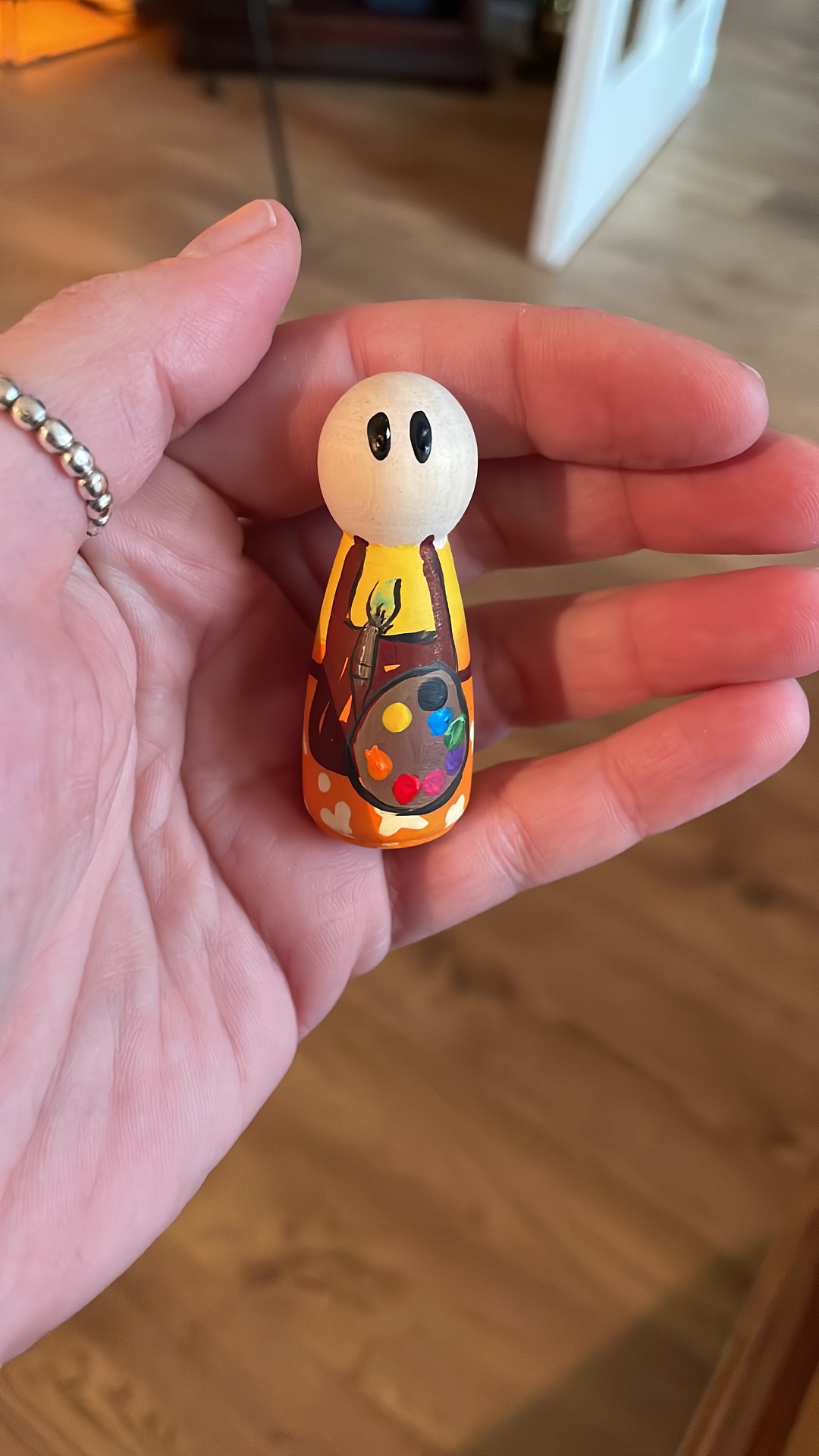 Hand houdt een kleine houten poppetje met wit hoofd, zwarte ogen en kleurrijke jas met hartjes. Handgeschilderd